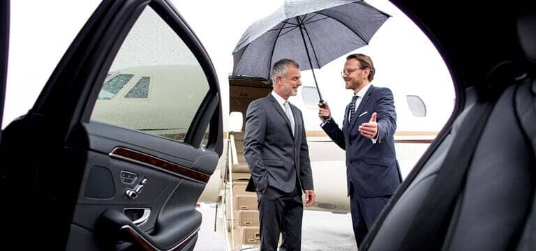 Two men in suits standing next to a car with an umbrella.